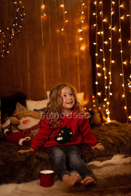 Очаровательная маленькая девочка с красной кружкой сидит в комнате, полной рождественских украшений и огней — стоковое фото