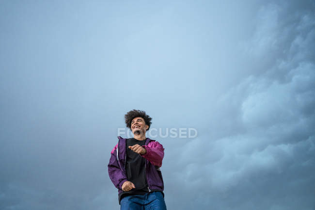 Снизу хипстера в стильной спортивной красочной одежде стоя и глядя в сторону с голубым небом на заднем плане — стоковое фото