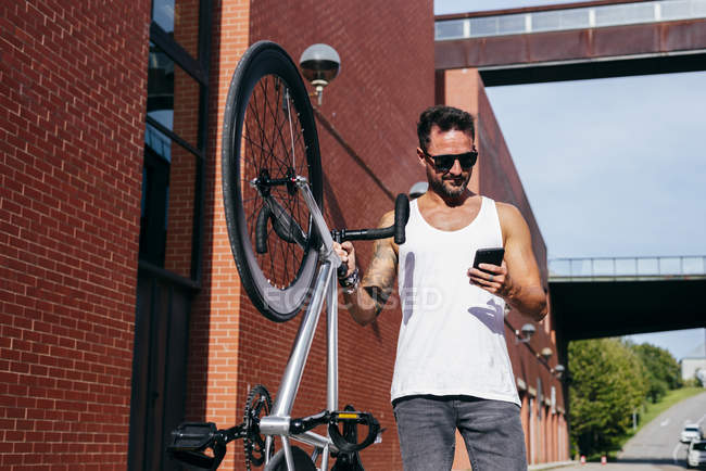 Guapo ciclista masculino en ropa deportiva y gafas de sol con teléfono inteligente mientras está de pie con la bicicleta junto a la pared de ladrillo rojo - foto de stock