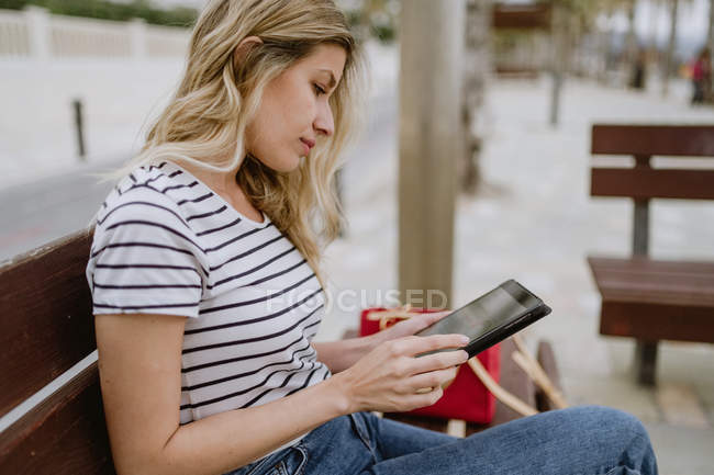 Frau in gestreiftem Hemd und Jeans sitzt mit Einwegkaffee auf einer Bank am Meer und surft mit Tablet im Internet — Stockfoto