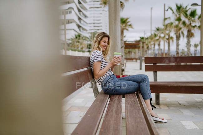 Vista lateral da mulher alegre bebendo de takeaway xícara de café sentado no banco da cidade em frente ao mar no dia de verão — Fotografia de Stock