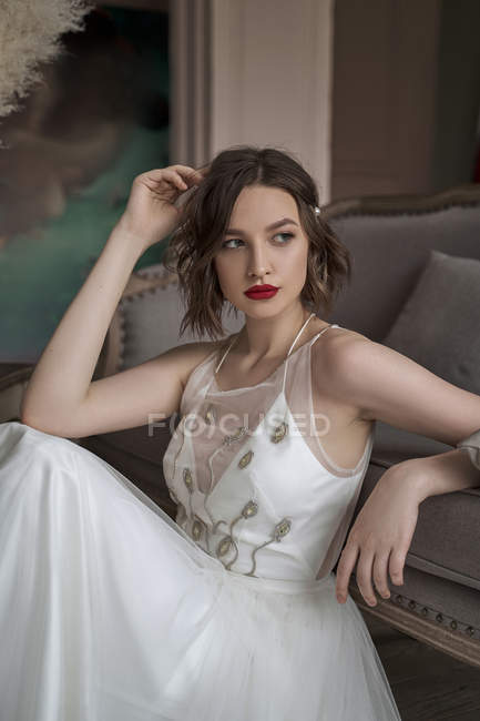 Hinreißende Frau mit roten Lippen im weißen Kleid, die wegschaut, während sie auf dem Boden neben dem Sofa sitzt — Stockfoto