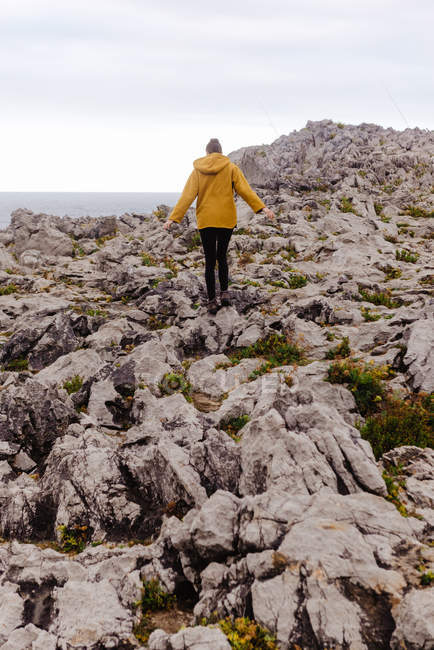 Vista posteriore della donna in felpa gialla con cappuccio che calpesta la riva pietrosa circondata da onde schiumose nella giornata nuvolosa grigia — Foto stock
