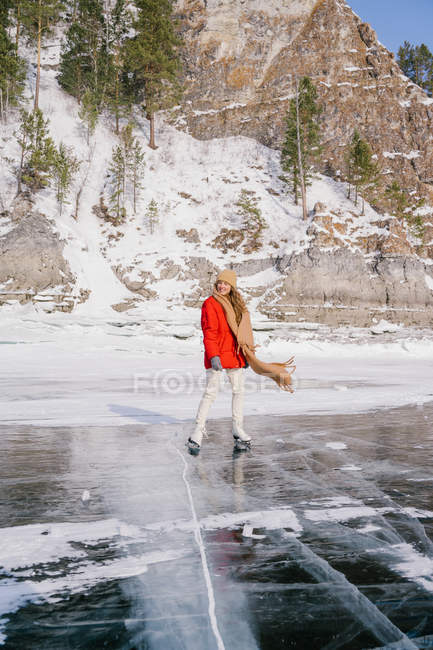 Happy active female in warm sportswear skating on ice on Siberian river next to rocks covered with snow and pine trees — Stock Photo