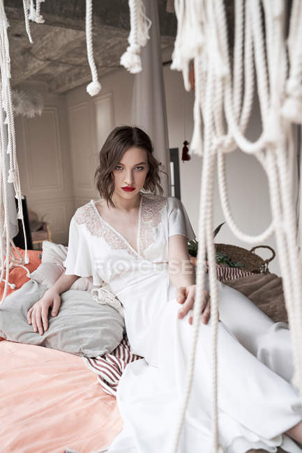 Preciosa mujer con labios rojos en vestido blanco mirando a la cámara mientras está sentada en el suelo junto al sofá - foto de stock