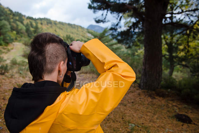Vista posteriore del giovane con acconciatura elegante scattare foto di albero nella valle di montagna in giornata grigia — Foto stock