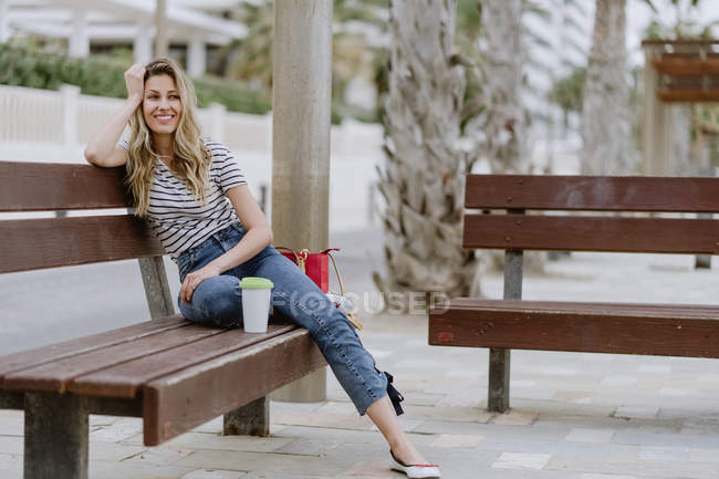 Vue latérale de la femme occasionnelle joyeuse assise sur le banc de la ville en bord de mer le jour d'été en regardant loin — Photo de stock
