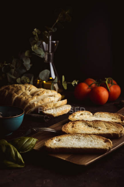 Шматочки свіжого хліба розміщені на столі біля свіжих помідорів і листя базиліка на чорному тлі — стокове фото