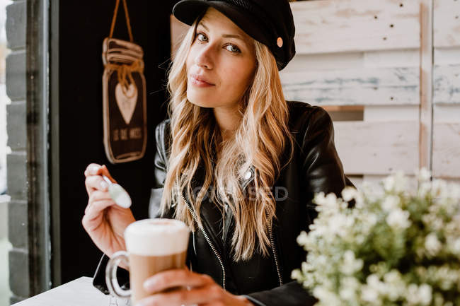 Довгошерста модна красива блондинка п'є зі склянки смачної пінистої кави, дивлячись на камеру — стокове фото