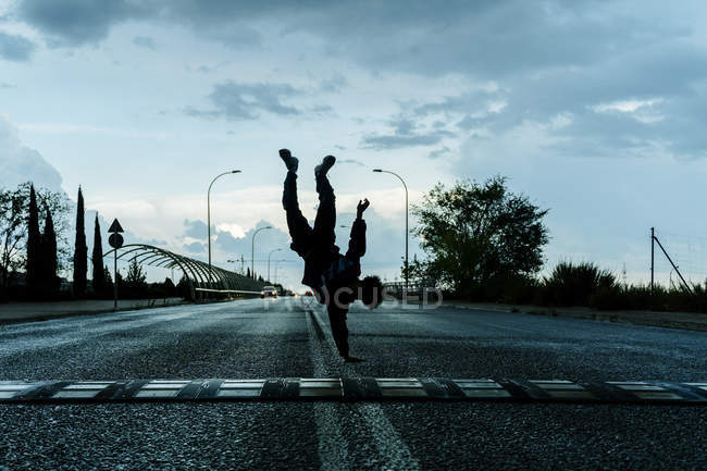 Silhouette of dancer in sportswear doing handstand amid empty road with cloudy sky and trees on background — Stock Photo