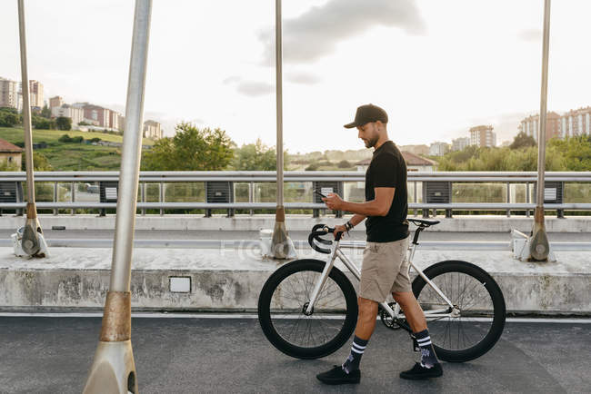 Beau cycliste masculin en vêtements de sport et lunettes de soleil à l'aide d'un smartphone tout en restant debout avec un vélo à côté du mur de briques rouges — Photo de stock