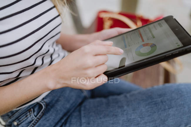 Cropped image of woman in striped shirt and jeans sitting on street bench browsing internet on tablet — Stock Photo