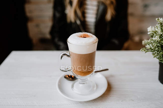 Cropped image of trendy woman sitting at table with glass of delicious foamy coffee on tabletop — Stock Photo