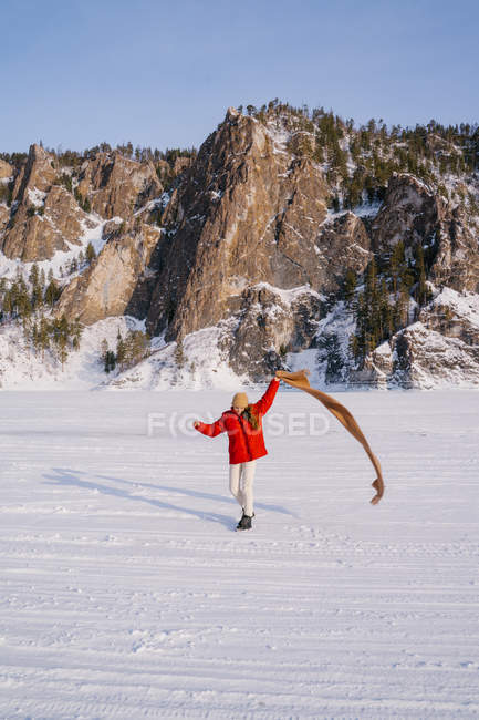 Glückliches Weibchen in roter Jacke läuft mit Schal in erhobener Hand auf schneebedecktem Tal mit majestätischen Felsen im Hintergrund — Stockfoto