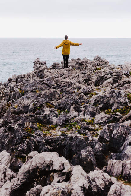 Visão traseira da mulher com capuz amarelo pisando na costa pedregosa cercada por ondas espumosas no dia nublado cinza — Fotografia de Stock