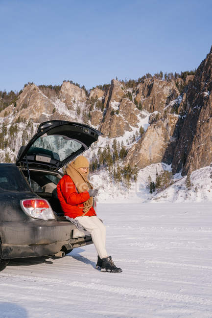 Side view of woman sitting on open car trunk and using phone in snowy valley with beautiful rocks during winter road trip — Stock Photo