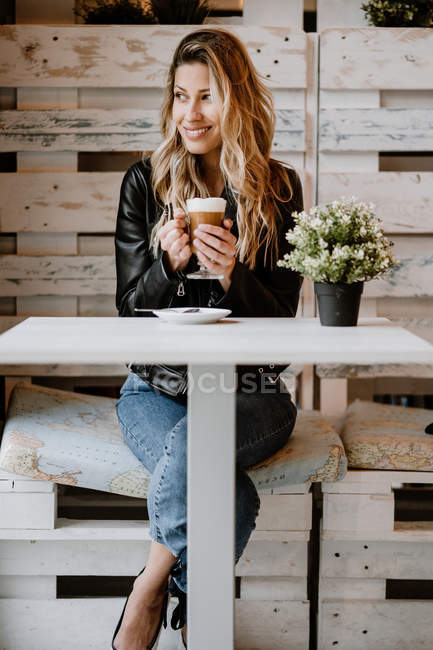 Long haired trendy beautiful blonde woman drinking from a glass of delicious foamy coffee while looking away — Stock Photo