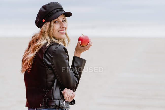 Vista trasera de la mujer rubia de moda en gorra negra y chaqueta de cuero mirando alegremente por encima del hombro con manzana roja madura en la mano en la playa de arena - foto de stock