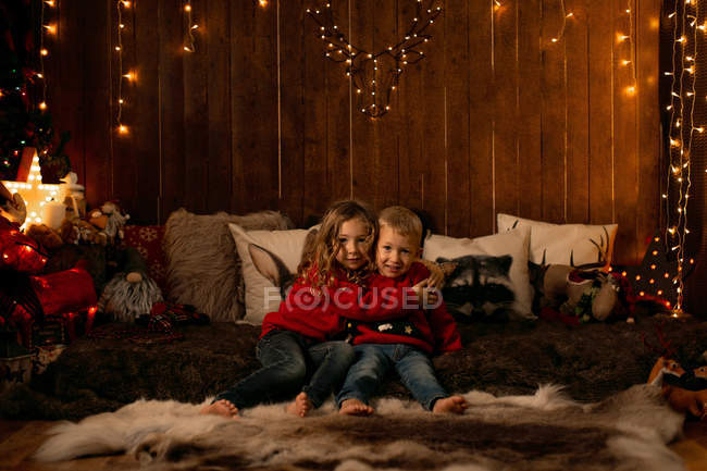 Очаровательная маленькая девочка и мальчик обнимаются, сидя в комнате, полной рождественских украшений, глядя в камеру — стоковое фото