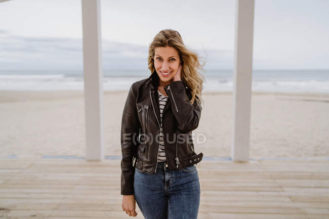 Trendfrau in schwarzer Lederjacke blickt selbstbewusst in die Kamera auf weißem Holzsteg mit Meer im Hintergrund — Stockfoto
