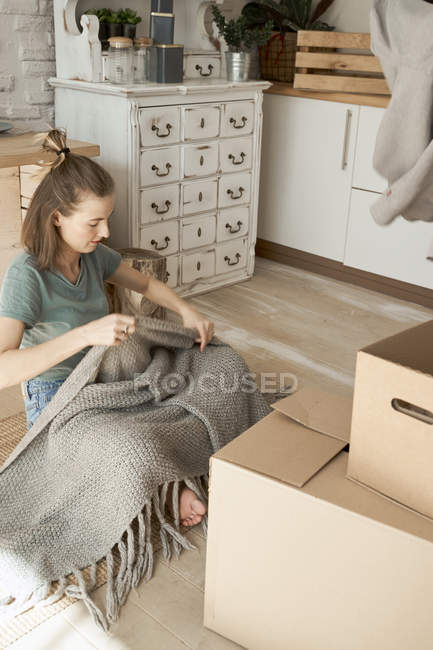 Молодая женщина достает мягкое серое одеяло из коробок в белой светлой кухне — стоковое фото