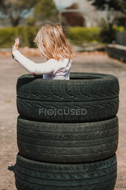 From Behind of little girl standing in stack of car tires while having fun and playing outdoors on summer day — Stock Photo