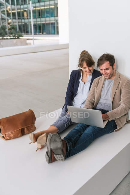 Задоволені бізнесмени посміхаються і переглядають ноутбук разом, сидячи за межами сучасної будівлі на міській вулиці — стокове фото