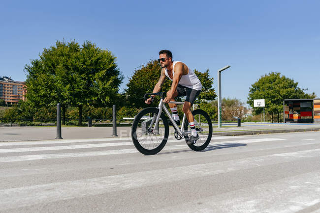 Спортсмен катается на велосипеде по городской дороге с зелеными деревьями на обочине дороги в летний день с голубым небом — стоковое фото