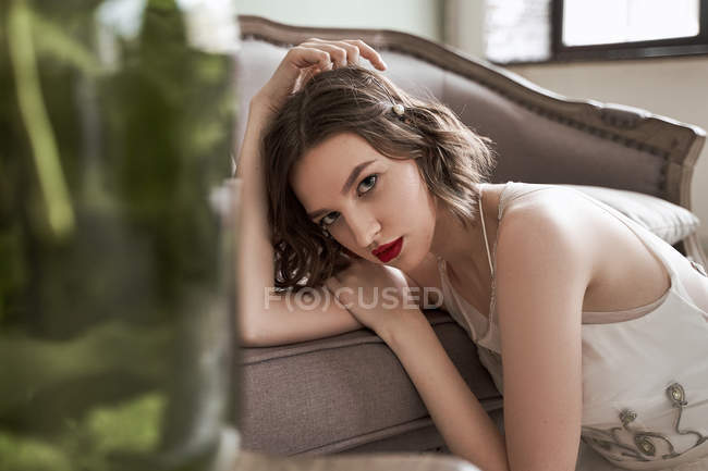Красива жінка з червоними губами в білому одязі дивиться на камеру, сидячи на підлозі біля дивана. — стокове фото