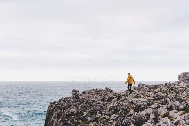 Side view of woman in yellow hoodie stepping on stony shore surrounded by foamy waves on gray cloudy day — Stock Photo