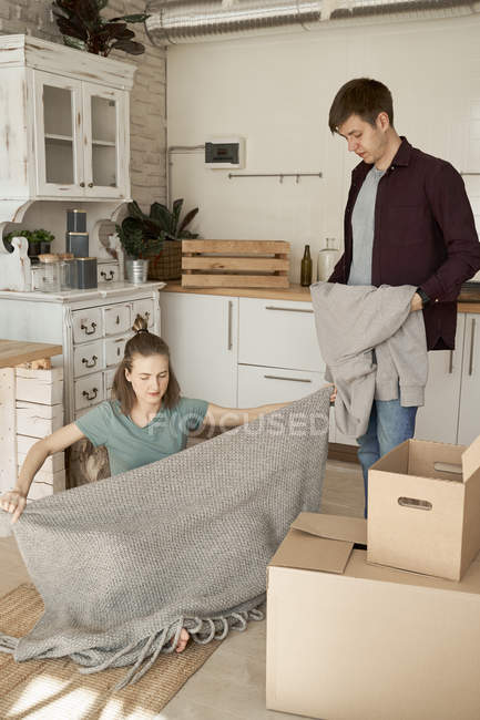 Happy young couple taking out soft gray sweater and blanket from boxes in kitchen — Stock Photo