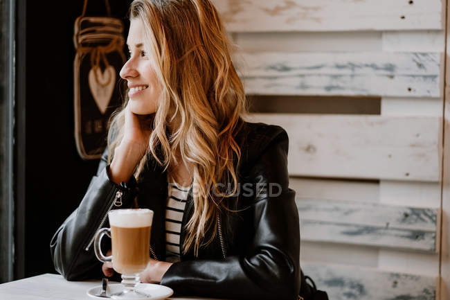 Vista lateral de la mujer rubia hermosa de moda de pelo largo sentada en una cafetería bebiendo de un vaso de delicioso café espumoso - foto de stock