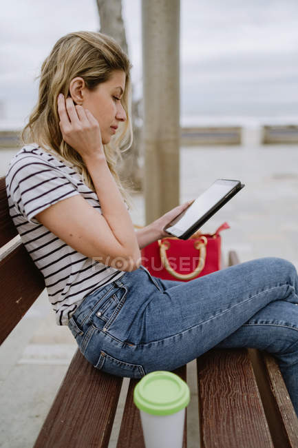 Vista lateral da mulher em camisa listrada e jeans sentado no banco de rua à beira-mar com xícara descartável de café e internet de navegação em tablet — Fotografia de Stock