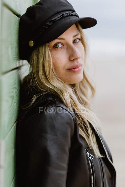 Frau in schwarzer Mütze und Lederjacke lächelt fröhlich in die Kamera und lehnt an Wand hölzerner Strandhütten — Stockfoto