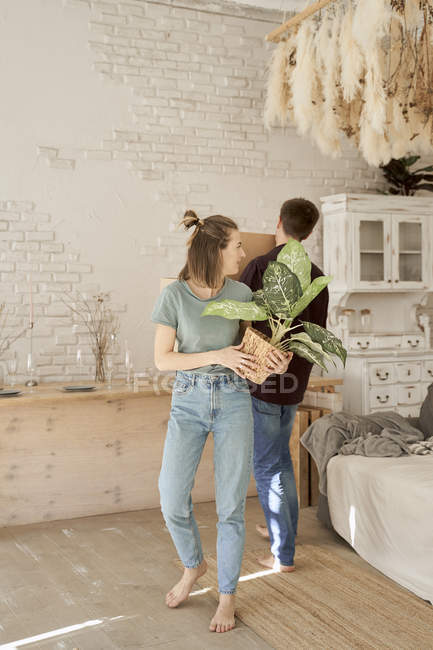Barefoot woman carrying pot with flower and looking at man in room furnishing new home — Stock Photo