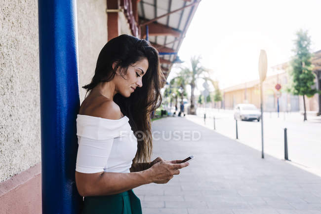 Joyeux jeune femme aux cheveux longs appuyé contre le mur en regardant le téléphone cellulaire — Photo de stock