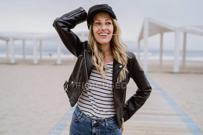 Trendy woman in black leather jacket confidently looking away on white wooden pier with ocean on background — Stock Photo