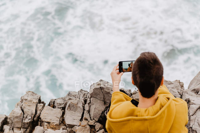 Rückansicht einer kurzhaarigen Frau in gelbem Sweatshirt, die am felsigen Meeresufer sitzt und ein Selfie mit dem Handy macht — Stockfoto