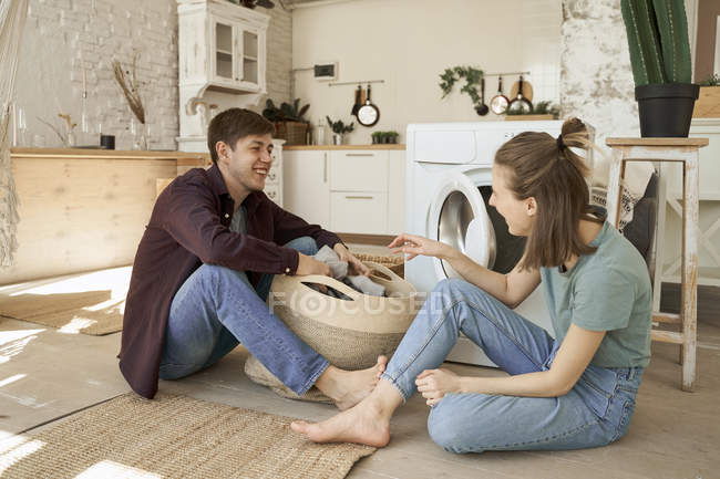 Happy young couple taking out clothes from basket in white light kitchen to put on washing machine — Stock Photo