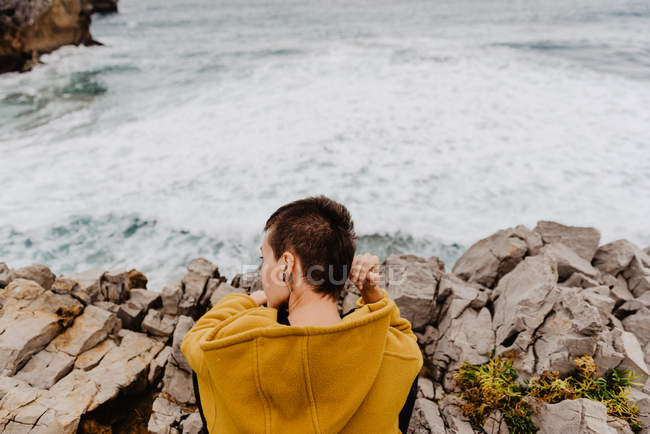Visão traseira da mulher com capuz quente amarelo sentado sozinho na costa rochosa olhando para ondas espumosas no dia nublado — Fotografia de Stock