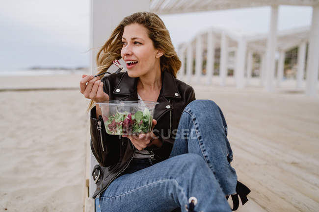 Стильна жінка в чорній куртці насолоджується здоровим зеленим салатом, сидячи на дерев'яній терасі на узбережжі, посміхаючись, дивлячись — стокове фото