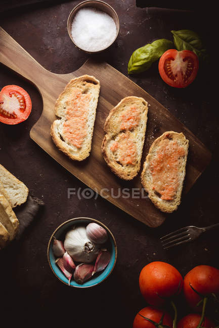 Spices, garlic and tomatoes near toasts on wooden board — Stock Photo