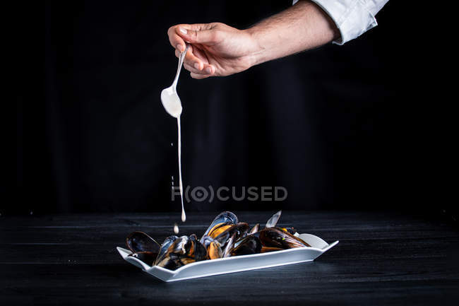 Crop cook pouring fragrant appetizing mussels with Thai sauce on black background — Stock Photo
