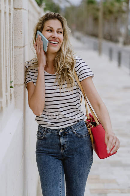 Щаслива жінка у смугастій сорочці й джинсах стоїть біля міської вулиці й розмовляє на смартфоні. — стокове фото