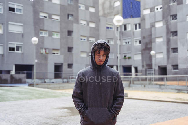 Pensive man in hoodie standing with eyes down with city buildings on background at cold day — Stock Photo