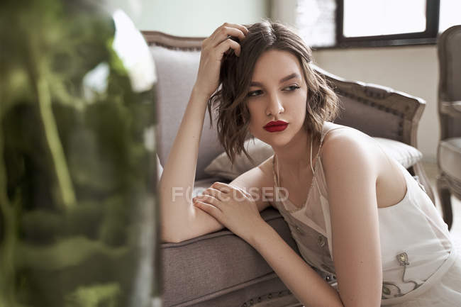 Gorgeous woman with red lips in white dress looking away while sitting on floor beside sofa — Stock Photo