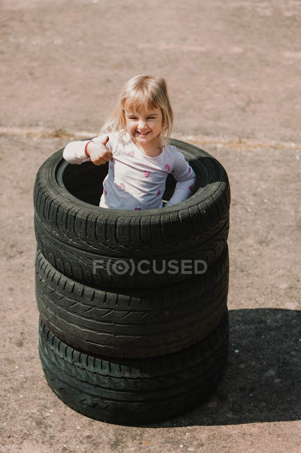 Зверху щаслива чарівна дівчинка, що стоїть в стосі автомобільних шин, роблячи знак ОК, розважаючись і граючи на відкритому повітрі в літній день — стокове фото