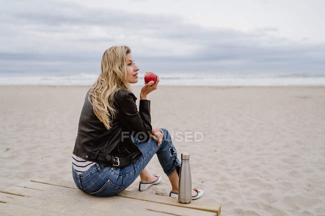 Vista laterale della donna alla moda in berretto nero e giacca di pelle mangiare mela rossa matura sulla spiaggia di sabbia — Foto stock