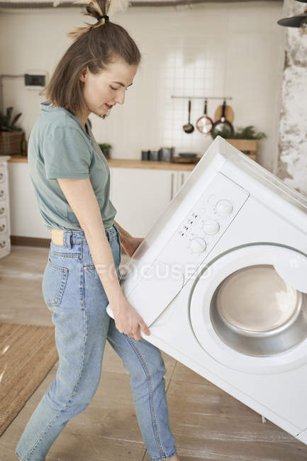 Мощная веселая молодая женщина пытается нести белую стиральную машину в одиночку на светлой кухне — стоковое фото