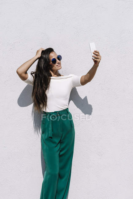 Приваблива молода жінка в модному вбранні позує за селфі, стоячи на яскравому фоні — стокове фото